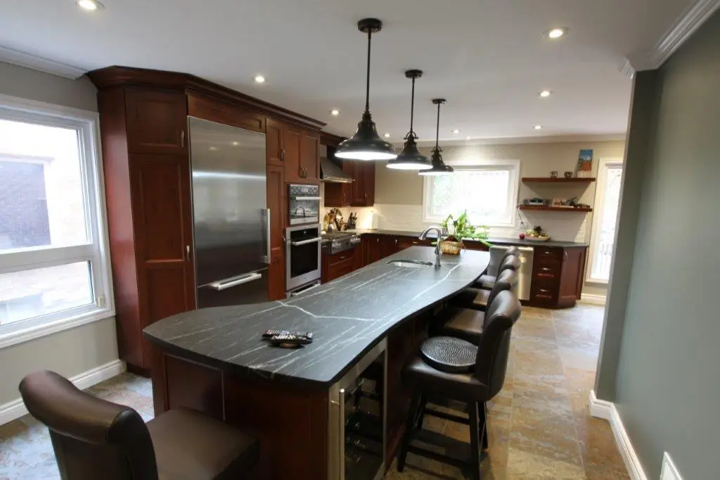Modern kitchen with a long black marble island countertop, dark brown wooden cabinets, stainless steel refrigerator and oven, four black cushioned bar stools, pendant lights hanging above the island, and large windows providing natural light.