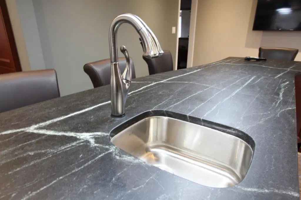 Close-up of a modern kitchen sink with a stainless steel faucet set into a black marble countertop with white veining, surrounded by brown chairs in a dining or kitchen area.