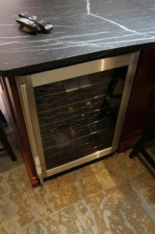 Built-in wine cooler with a glass door housing several wine bottles, set beneath a black marble countertop with white veins, and a few remote controls placed on the countertop.
