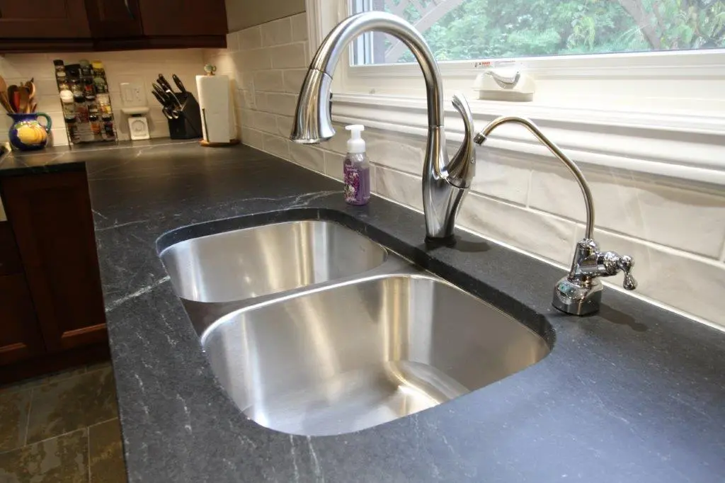 Modern double stainless steel kitchen sink with tall curved faucet and separate water filter faucet on a dark gray stone countertop, surrounded by wooden cabinets and a white tiled backsplash beneath a window.
