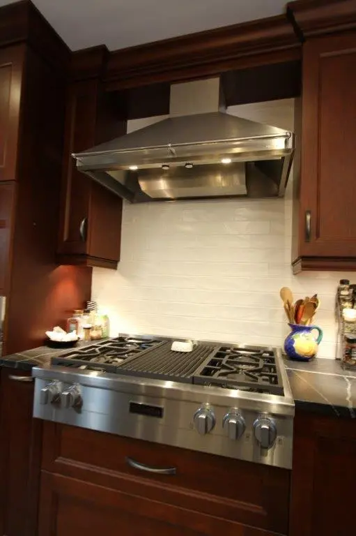 Modern kitchen stove with stainless steel range hood, surrounded by dark wooden cabinets and white subway tile backsplash, with cooking utensils in a blue and yellow ceramic holder on the right and jars on the left.