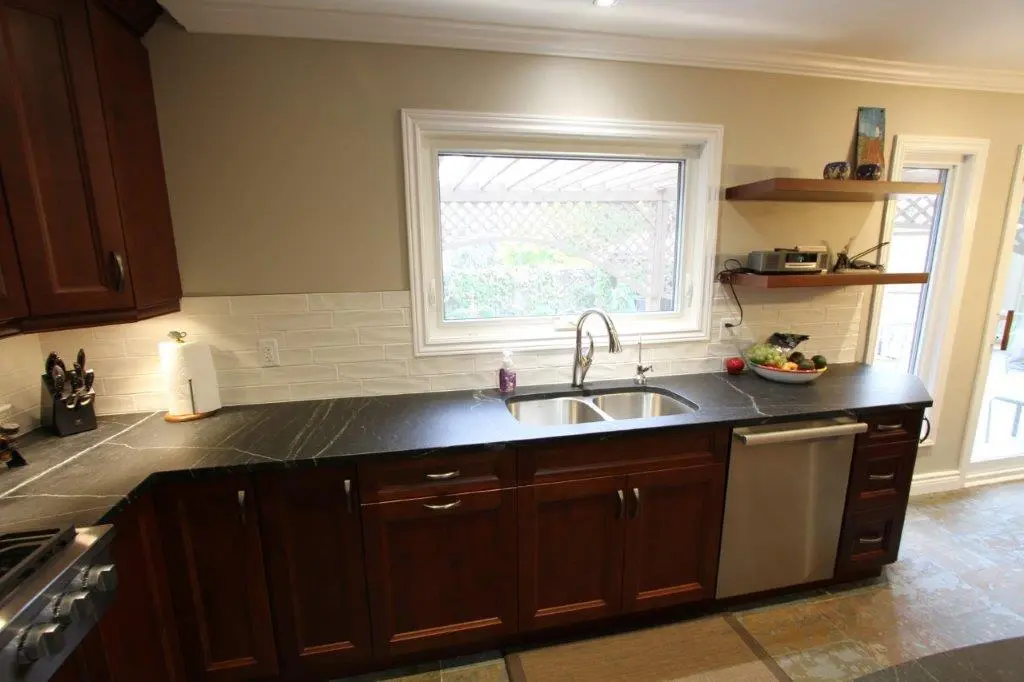 Modern kitchen countertop with black surface, dual stainless steel sink under a window, dark wooden cabinets, a dishwasher, and shelves holding a stereo and fruit bowl.