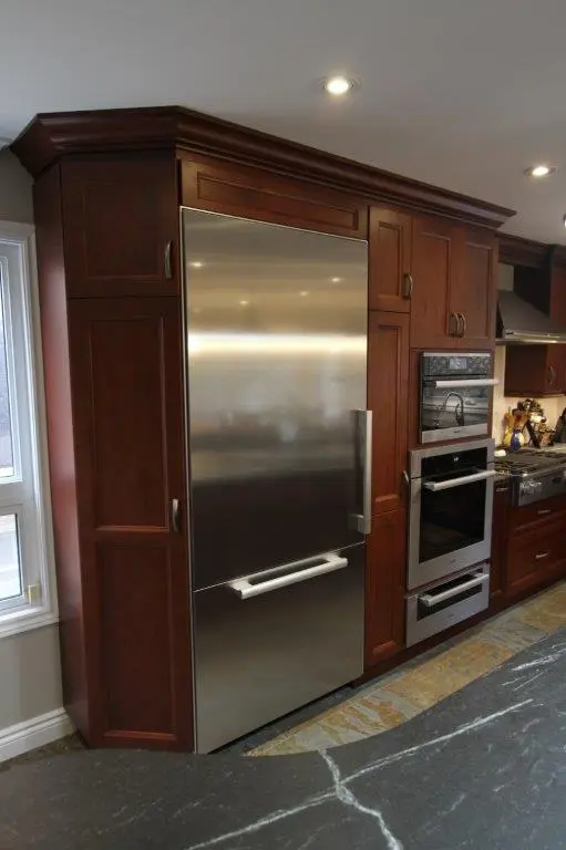 Built-in stainless steel refrigerator integrated with dark wooden cabinets in a modern kitchen featuring an oven and a gas stove with a dark granite countertop in the foreground.