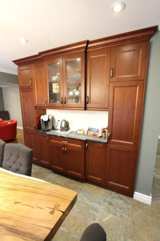 Built-in dark wood kitchen cabinets with glass window doors on upper section, countertop with coffee maker and kettle, and surrounding cushioned dining chairs.