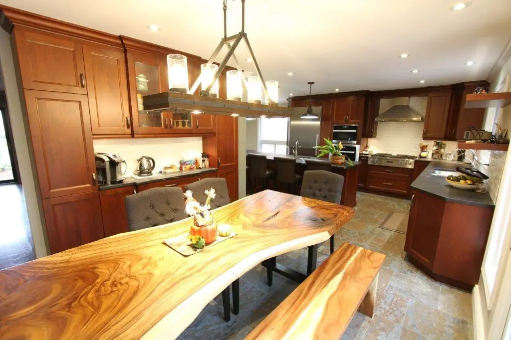 Modern kitchen with dark wooden cabinets, a natural wood live-edge dining table and matching bench, gray upholstered chairs, stone tile floor, and hanging rectangular light fixture with glass shades.