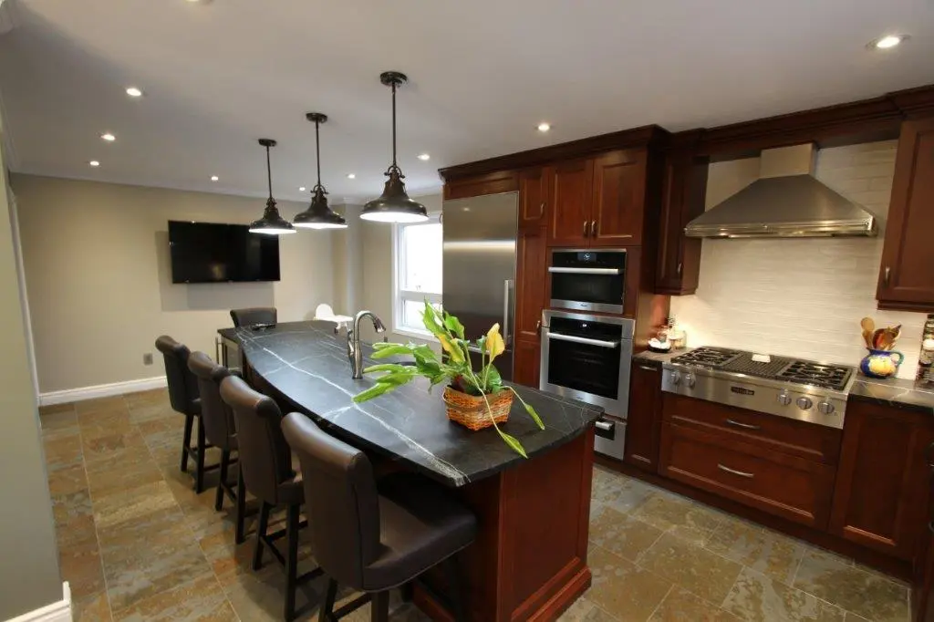 Modern kitchen with dark wood cabinets, stainless steel appliances, a black stone countertop island with brown cushioned chairs, three pendant lights overhead, a potted plant centerpiece, and a tiled floor.