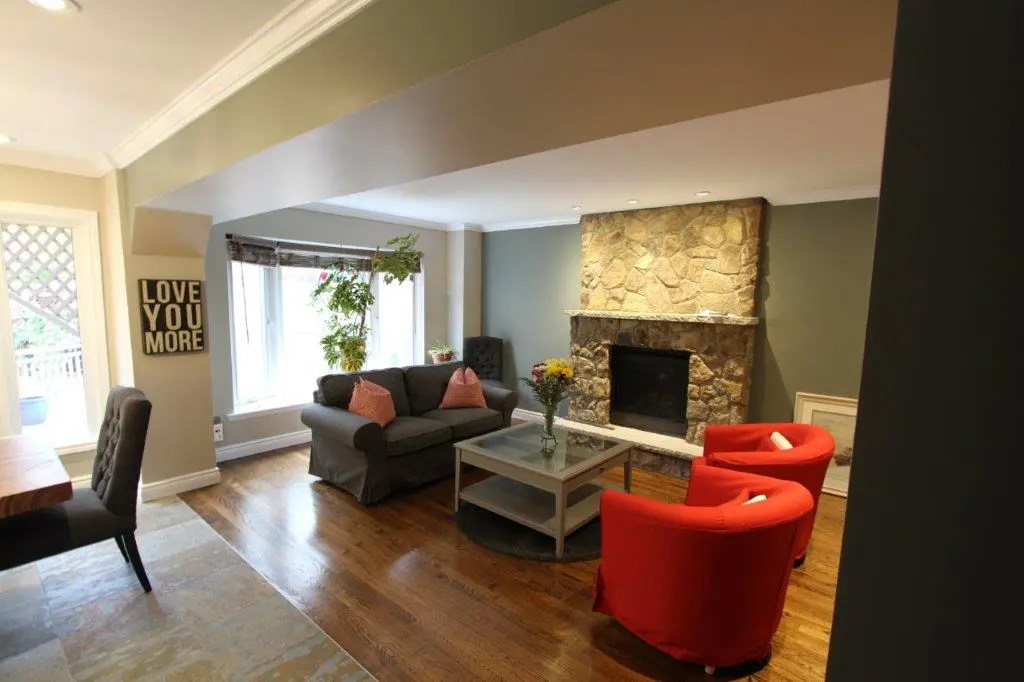 Cozy living room with a stone fireplace, a gray sofa with pink pillows, two bright red armchairs, a glass coffee table with flowers, wooden floor, and large windows with plants, along with a 'LOVE YOU MORE' sign on the wall near the entrance.