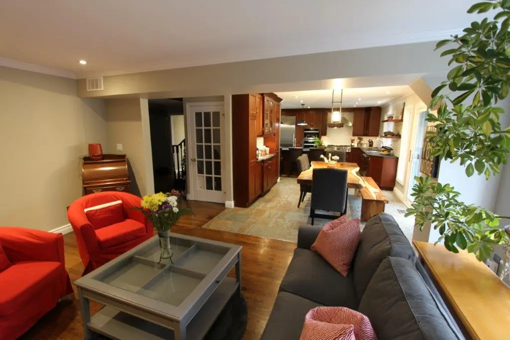 Open living room and kitchen area with gray sofa and red cushions, two red armchairs, glass coffee table with flowers, wooden floor, wooden dining table with chairs, dark wood kitchen cabinets, and green plant near the window.