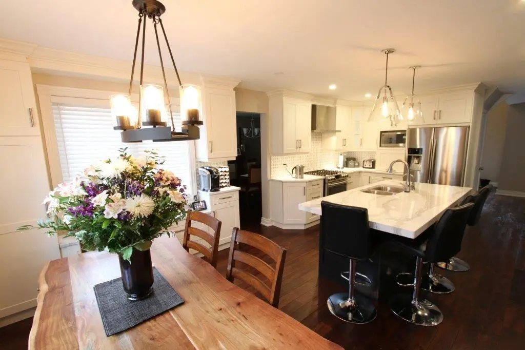 Bright modern kitchen with white cabinetry, a large island with a white countertop and three black swivel stools, wooden dining table with a large floral arrangement in a dark vase, hardwood flooring, and stainless steel appliances including a fridge and oven.