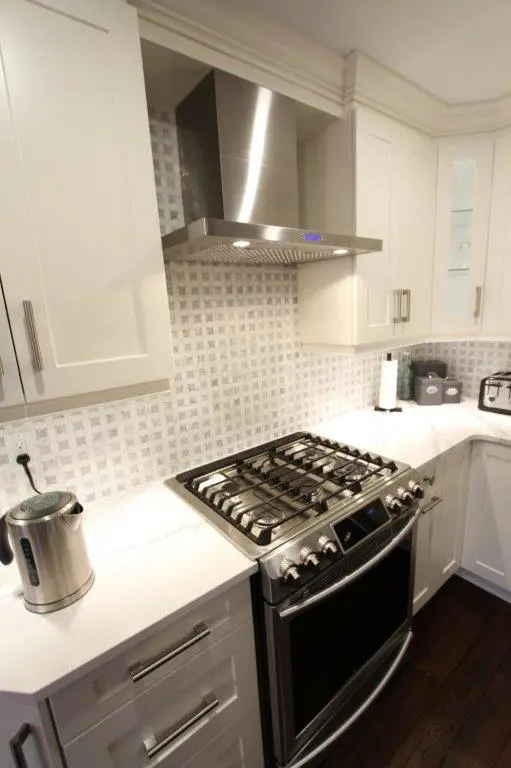 Modern kitchen corner with white cabinets, a stainless steel gas stove and oven, a stainless steel range hood, a kettle on the counter, and a light gray patterned backsplash.