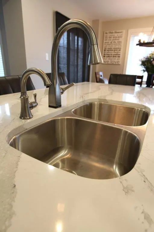 Double stainless steel kitchen sink with brushed nickel faucets set in a white marble countertop in a modern kitchen dining area.