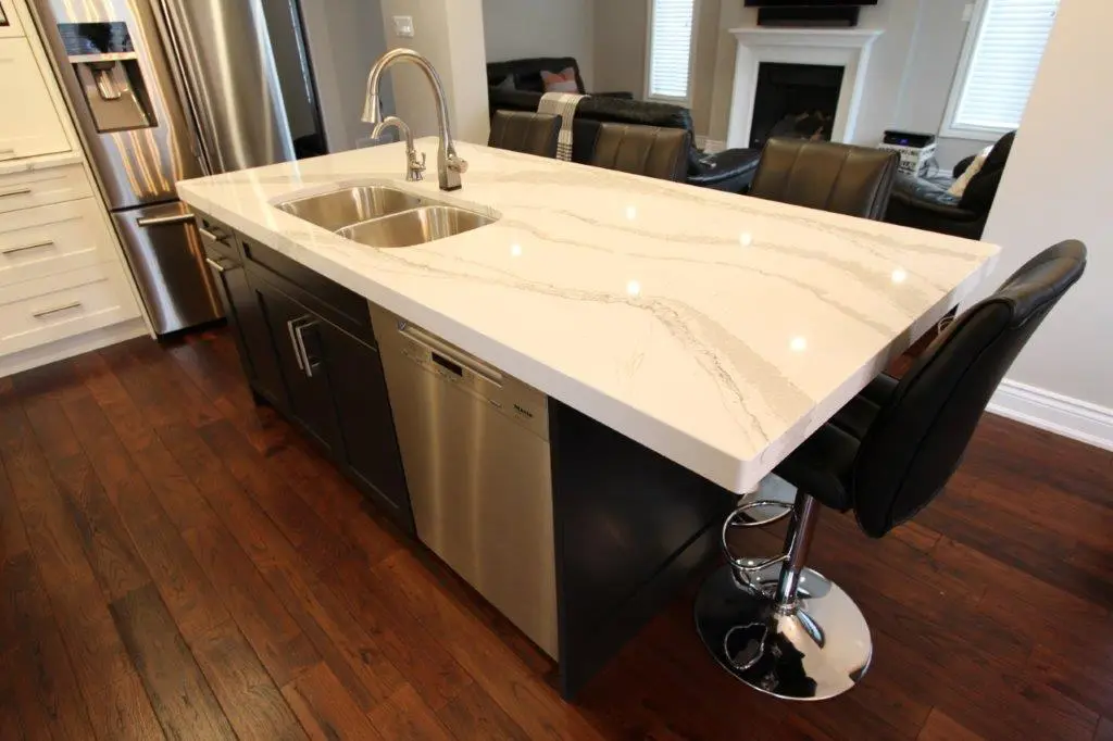 Modern kitchen island with a white marble countertop, double stainless steel sink, and a chrome faucet, surrounded by black leather bar stools, with dark wooden flooring and stainless steel appliances nearby.