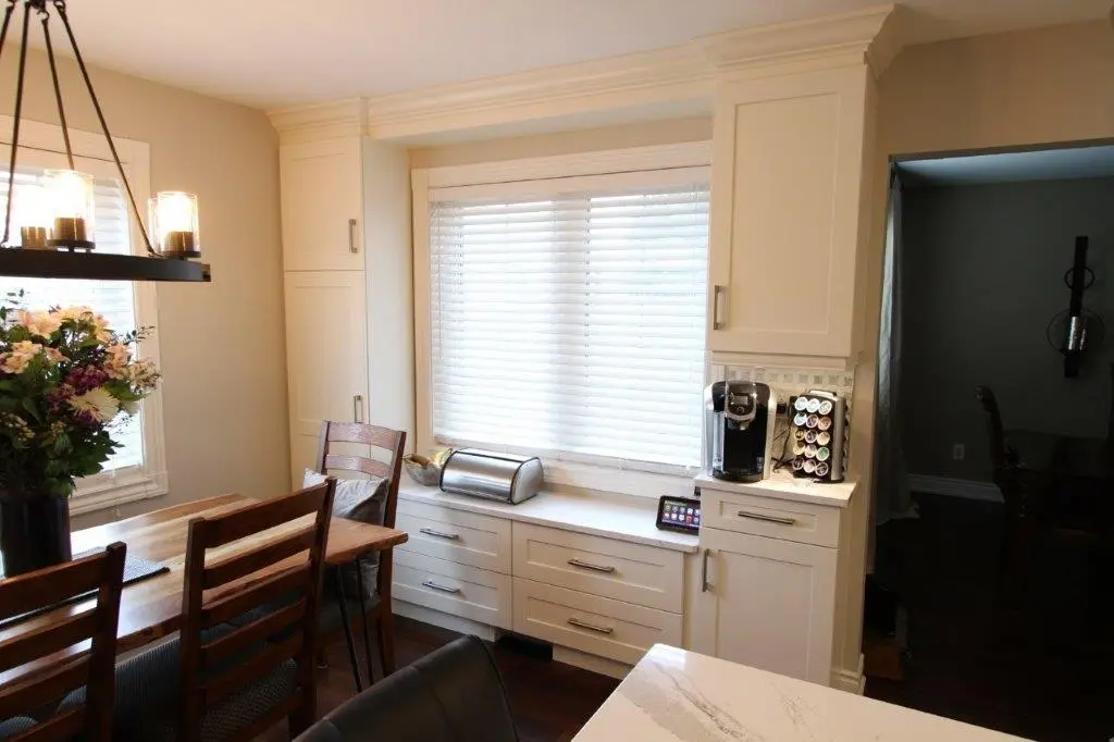 Kitchen area with white cabinetry and a large window with blinds above drawers, a coffee maker with a pod holder on the counter, a black metal chandelier with glass shades above a wooden dining table with chairs and a vase of flowers, and a marble countertop in the foreground.