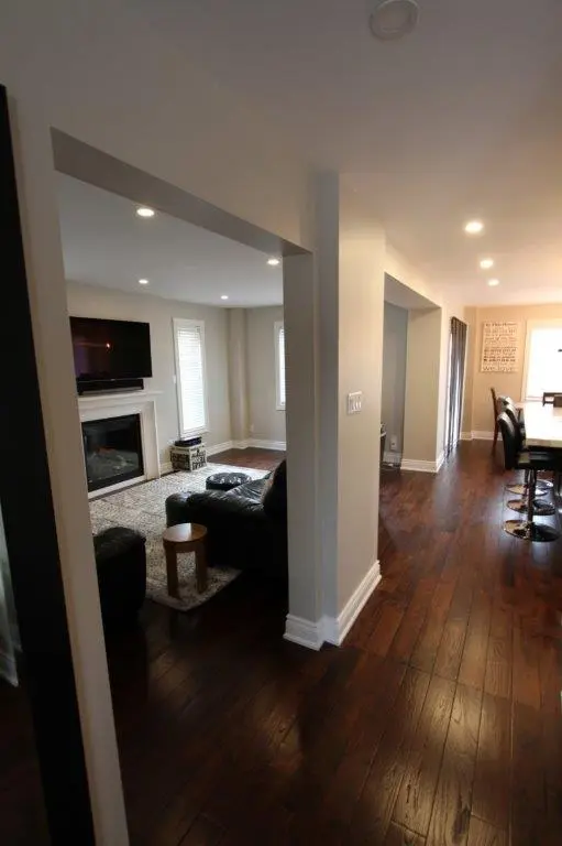 Interior view of a modern home with dark hardwood floors and beige walls, showing a living room with a wall-mounted TV above a white fireplace, black leather furniture, a small wooden side table, and a carpet, as well as a hallway leading to a dining area with bar stools and a large window.