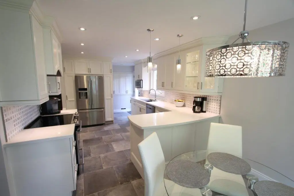 Modern kitchen with white cabinetry, stainless steel refrigerator and appliances, gray tiled floor, glass dining table with four white chairs, and decorative pendant lighting fixtures.