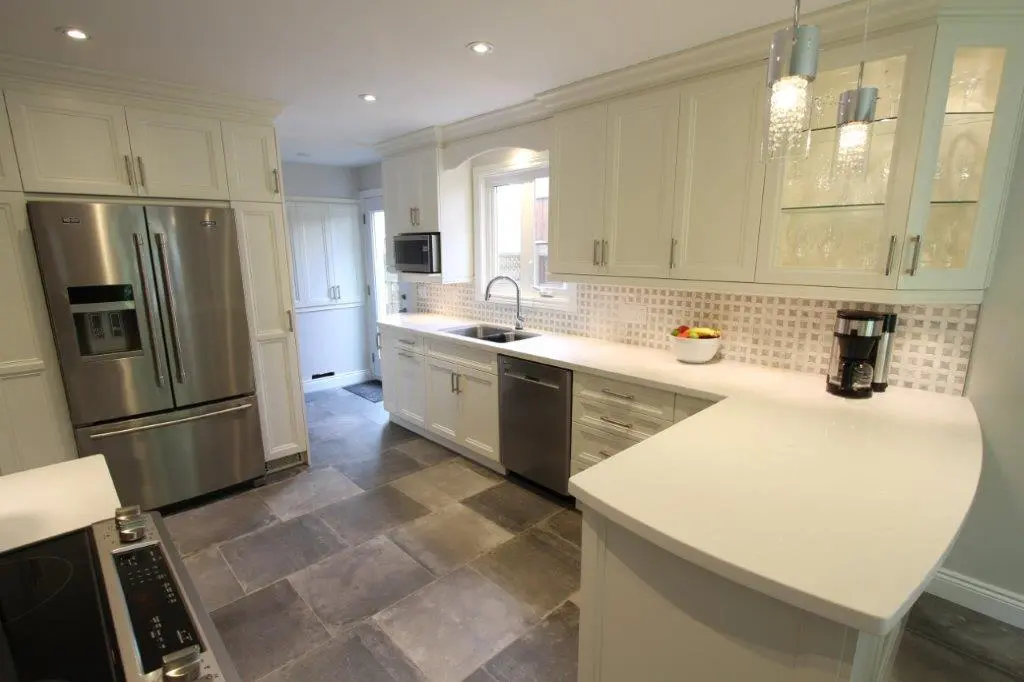 Modern kitchen with white cabinetry, stainless steel refrigerator, dishwasher, microwave, and a coffee maker on a white countertop under glass-front cabinets, with tiled floor and backsplash.