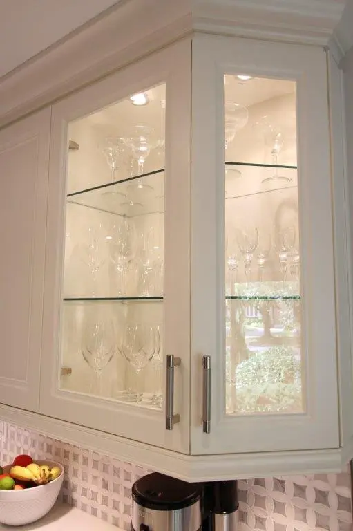 White kitchen corner cabinet with glass doors displaying various clear wine glasses on glass shelves, with a white countertop below holding a bowl of mixed fruit and a black appliance against a patterned backsplash.