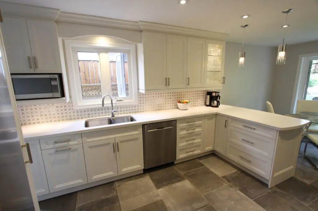 Bright modern kitchen with white cabinets, stainless steel sink and dishwasher, a microwave, coffee maker, and a bowl of fruit on the counter, with a tiled backsplash and large gray floor tiles