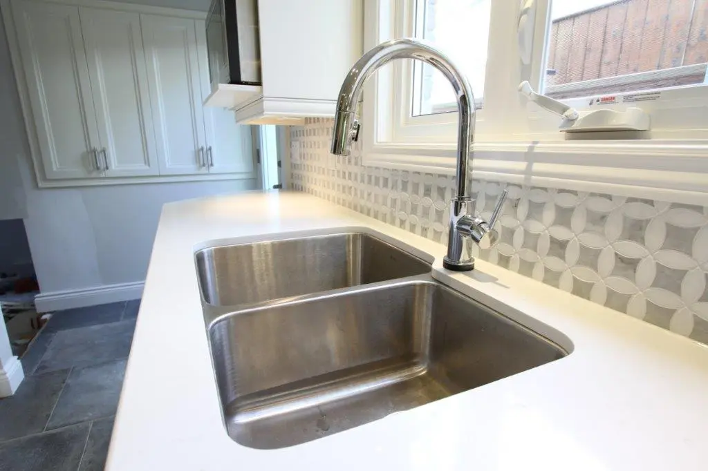 Modern kitchen countertop with a double stainless steel sink and a sleek chrome faucet beneath a window with patterned tile backsplash.