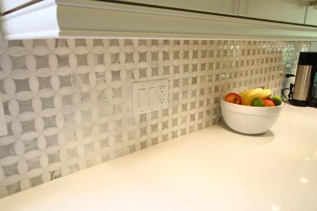 Kitchen countertop with a white and gray circular patterned tile backsplash, a white bowl filled with bananas, apples, and limes, and a coffee maker on the right side.