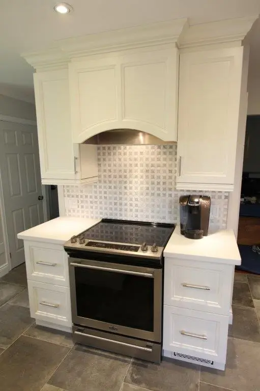 Modern kitchen area with a black and stainless steel electric stove and oven, surrounded by white cabinetry and countertops, with a silver coffee machine on the right counter and a tiled backsplash in a square pattern.