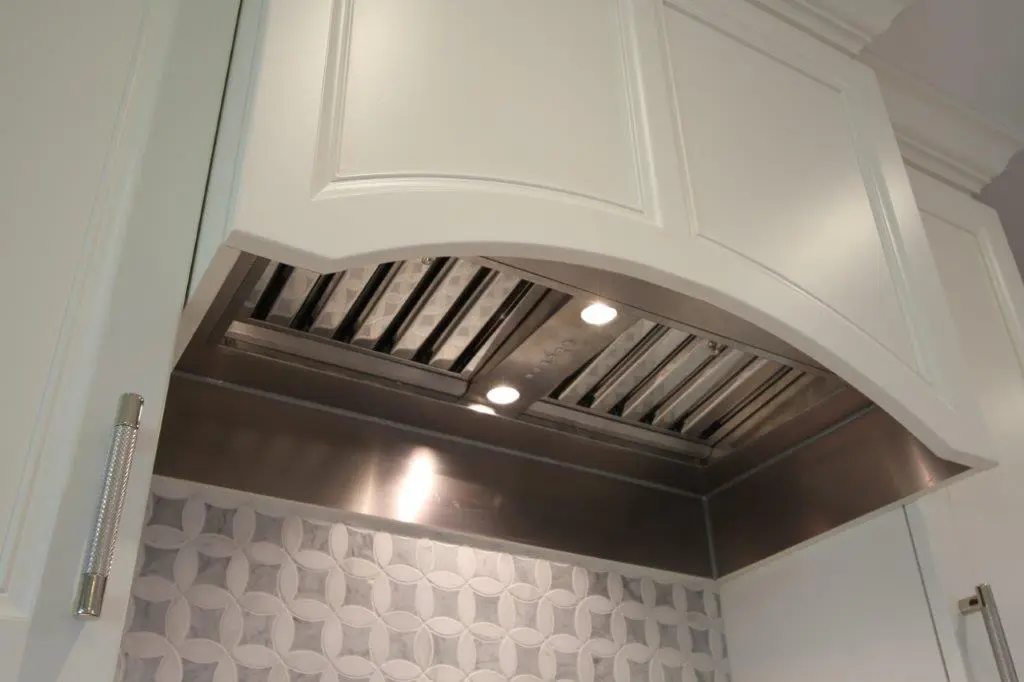 Stainless steel kitchen range hood with built-in lights mounted under a white cabinet above a patterned tile backsplash.