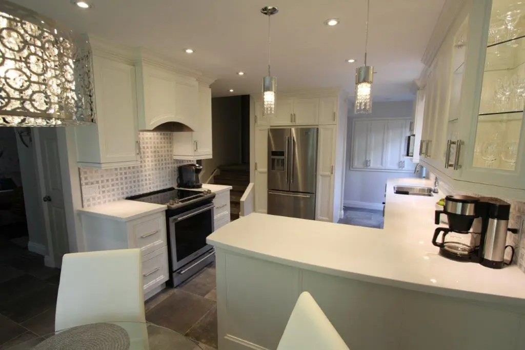 Modern kitchen with white cabinets, stainless steel refrigerator and oven, a white countertop with a coffee maker, and grey tiled floor with recessed and pendant lighting.