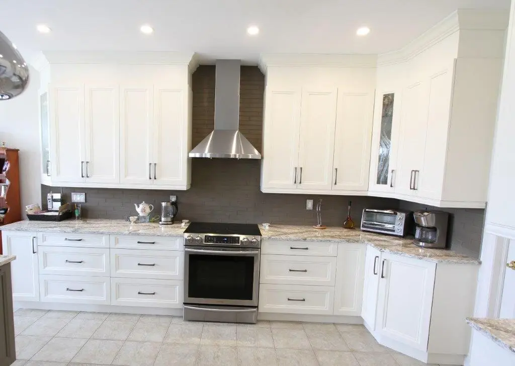 Modern kitchen with white cabinets, granite countertops, gray tile backsplash, stainless steel oven and range hood, coffee maker, and toaster oven on countertops.