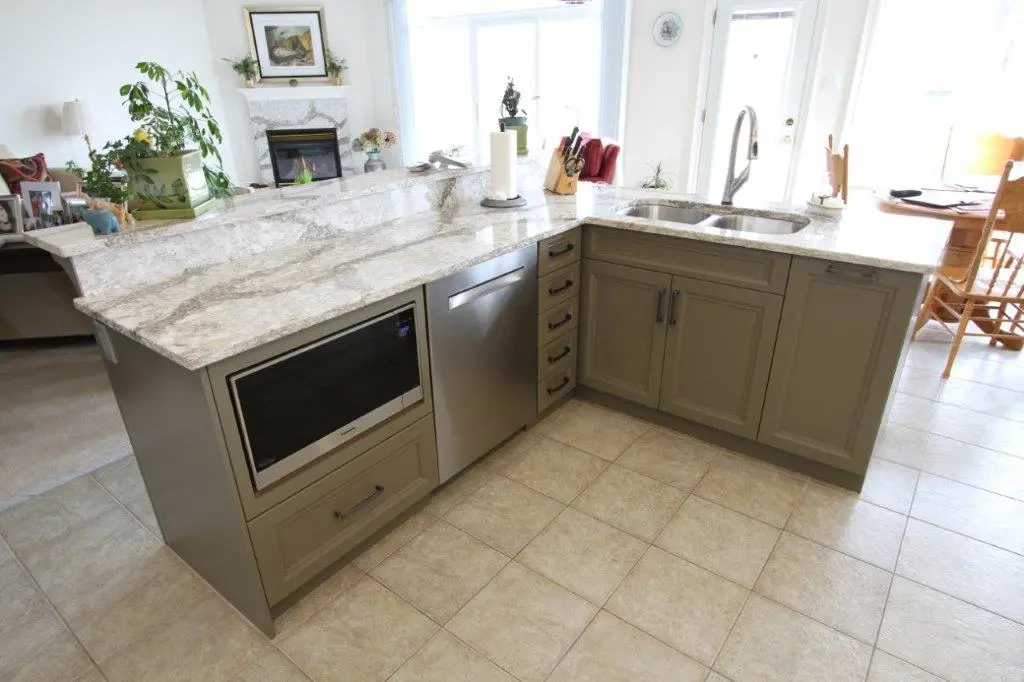 Modern kitchen island with beige cabinets and drawers, built-in microwave and dishwasher, granite countertop with paper towel holder, and double sink with a tall faucet, surrounded by light tile floor and dining area in the background.