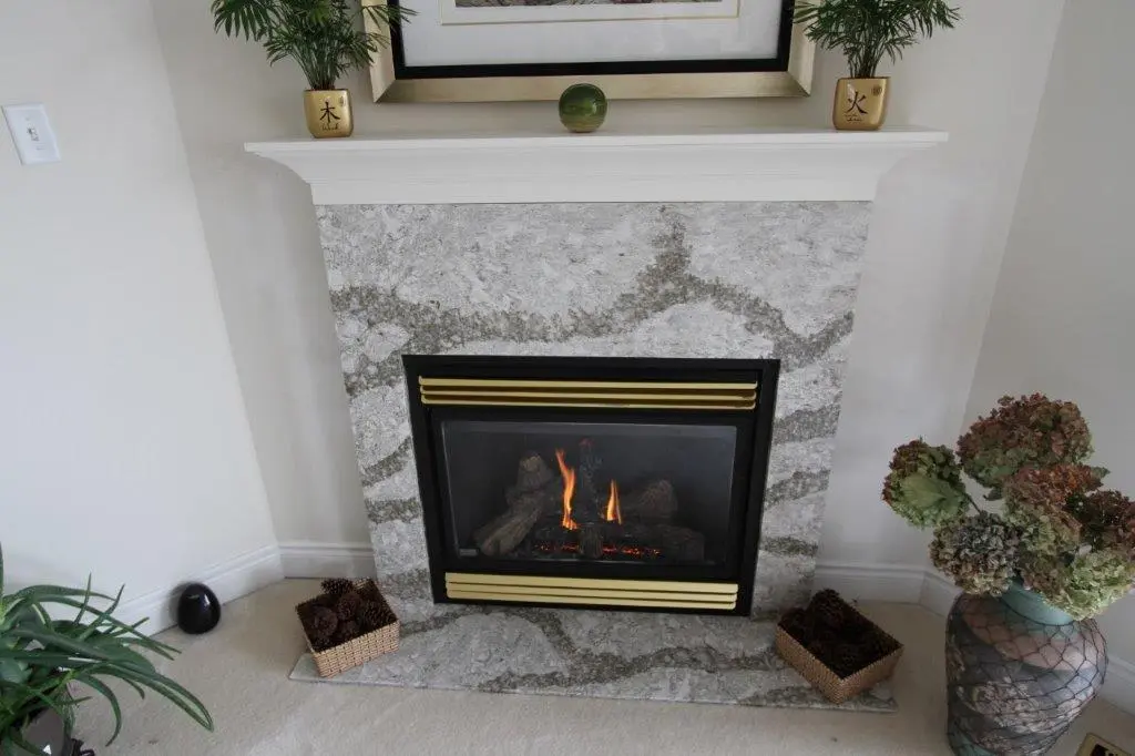 Modern fireplace with a marble surround and small flames inside, flanked by two small baskets with pine cones on the floor and plants on either side, topped with a white mantel holding two small gold pots with green plants and a small green spherical decoration in the center.