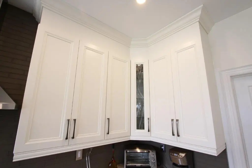 White kitchen corner cabinets with silver handles and a glass-fronted middle cabinet above a countertop with a toaster oven and coffee maker against a dark tiled backsplash.