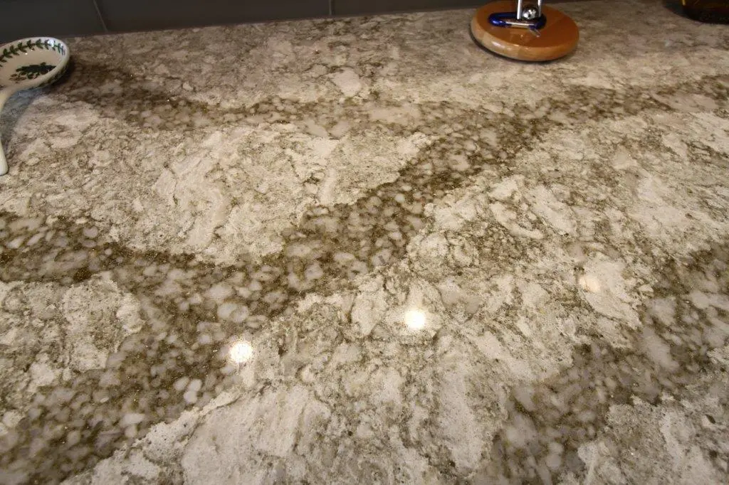 Close-up view of a polished granite countertop with white, gray, and brown veining and two reflections of round overhead lights, with a small decorative spoon and a wooden tape measure on the edges.