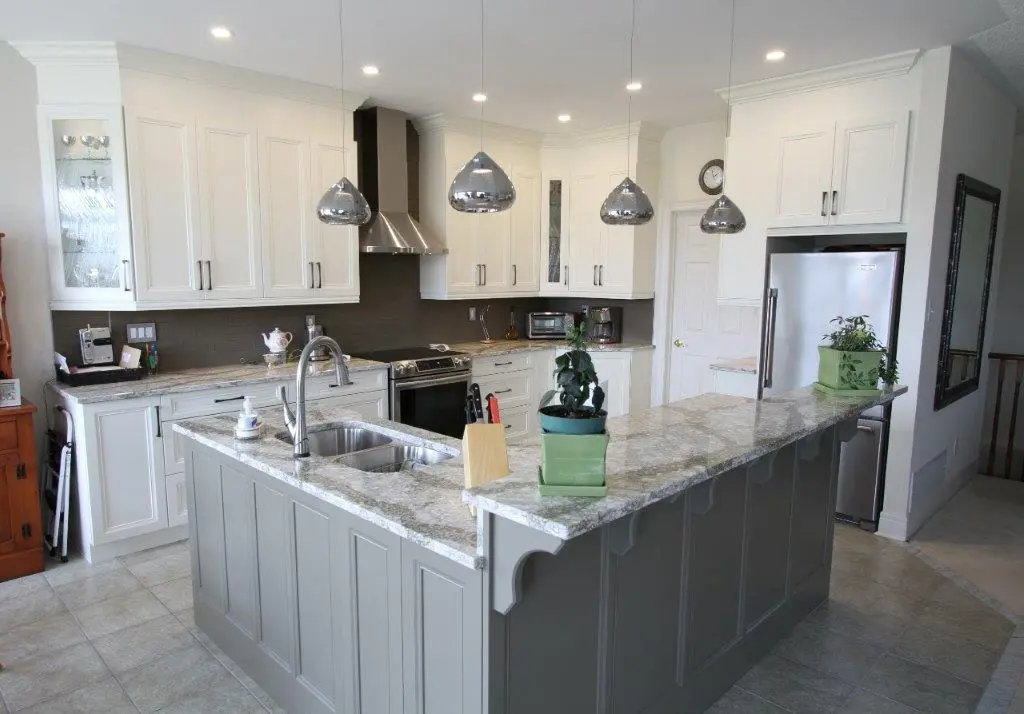 Modern kitchen with white upper cabinets and gray lower island cabinets, granite countertops, stainless steel appliances including stove and refrigerator, a double sink on the island, three silver pendant lights hanging over the island, and two small potted plants on the island countertop.