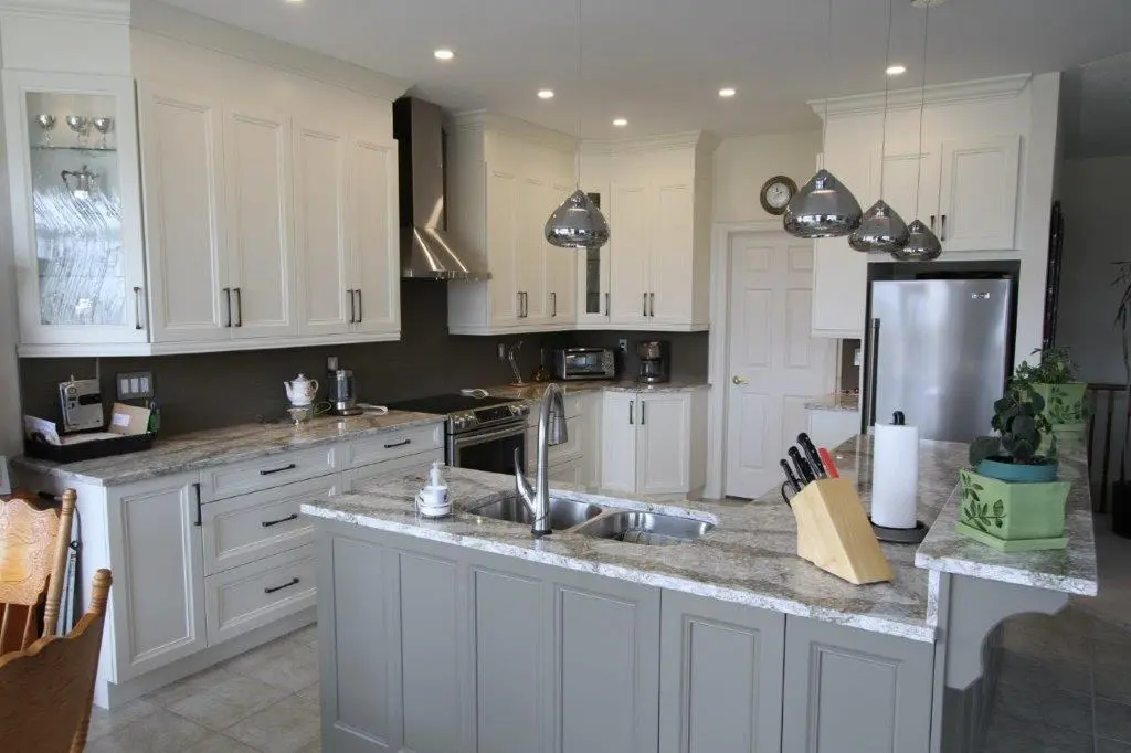 Modern kitchen with white cabinets, gray island with marble countertop, stainless steel double sink, stainless steel appliances, hanging pendant lights, and various kitchen items including a knife block, paper towel holder, and potted plants.