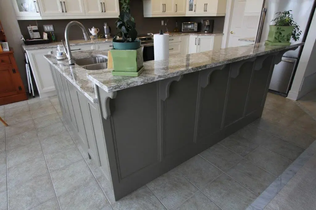 Modern kitchen island with gray cabinetry, granite countertop, stainless steel sink and faucet, two green potted plants, and a paper towel roll, surrounded by beige tiled flooring and white upper cabinets in the background
