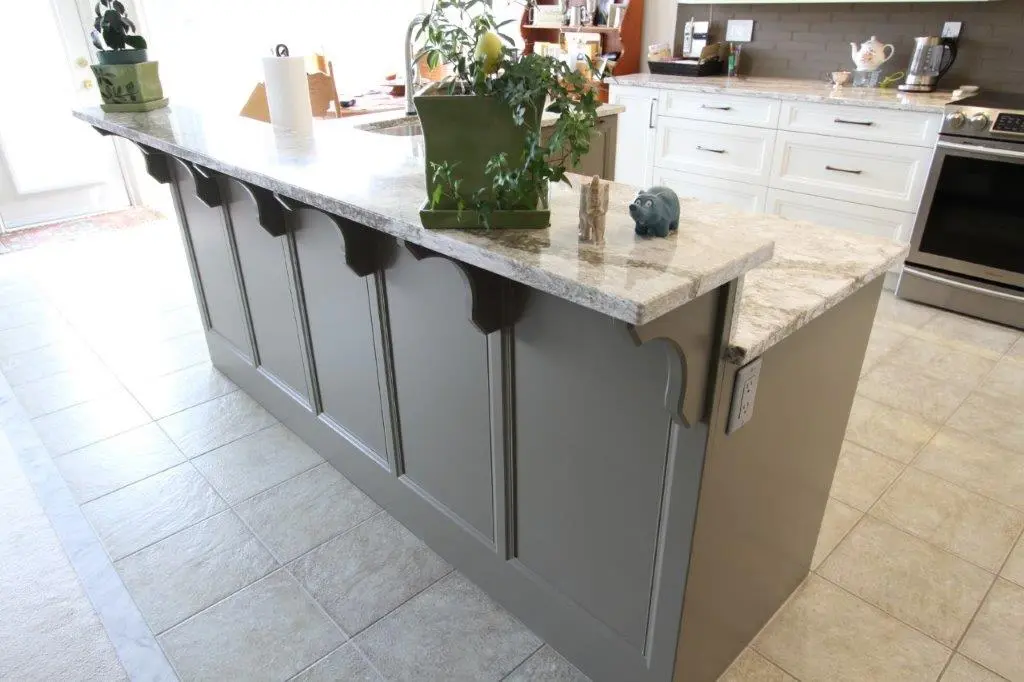 Kitchen island with gray paneled sides and a granite countertop, decorated with a green potted plant and two small animal figurines, set on a tiled floor with white kitchen cabinets and stainless steel appliances in the background.