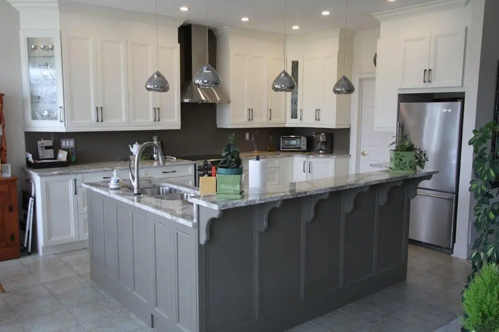 Modern kitchen with white upper cabinets, dark gray lower cabinets on the island, stainless steel refrigerator and hood, three hanging chrome pendant lights, granite countertops, and plants on the island and near the refrigerator.
