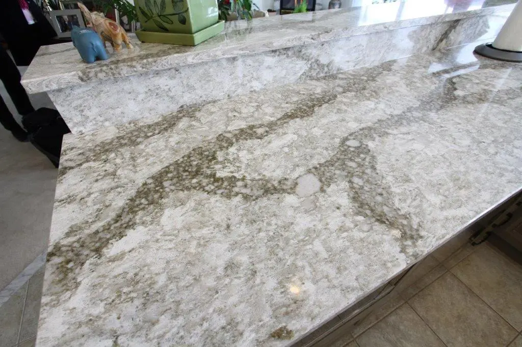 Close-up view of a light-colored granite kitchen countertop with natural brown and white patterns, part of a two-level counter setup with decorative items in the background and tiled floor visible.