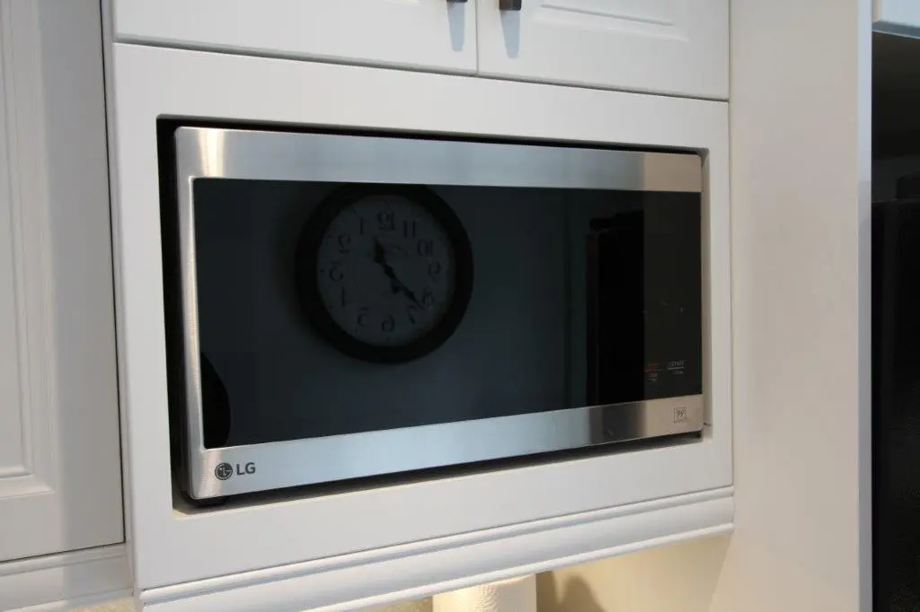 Built-in stainless steel LG microwave oven integrated into white kitchen cabinetry with a reflection of a wall clock on its glass door.