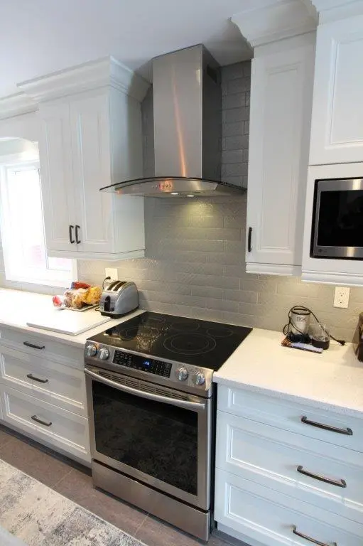 Modern kitchen with white cabinetry, stainless steel electric stove with black glass cooktop, stainless steel range hood, gray subway tile backsplash, white quartz countertops, toaster, and small kitchen appliances on the counter.