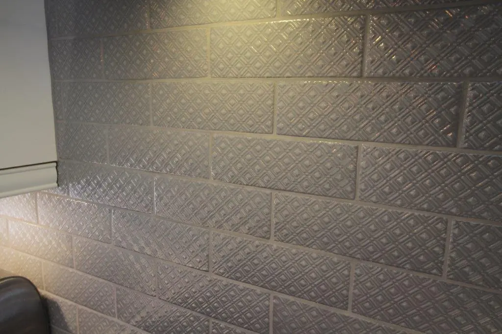 Gray ceramic tiles with a raised diamond pattern arranged in a staggered horizontal layout on a wall, partially illuminated by a warm light.