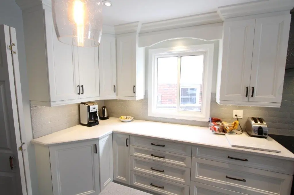 Bright kitchen corner with white cabinets, gray tiled backsplash, white countertops, a coffee maker, a kettle, a bowl, bags of bread, and a toaster near a window.