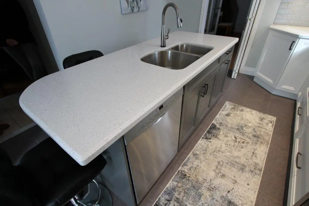 Modern kitchen island with a white speckled quartz countertop, stainless steel double sink, attached dishwasher, and gray cabinets, with black bar stools and a gray patterned rug on the floor.