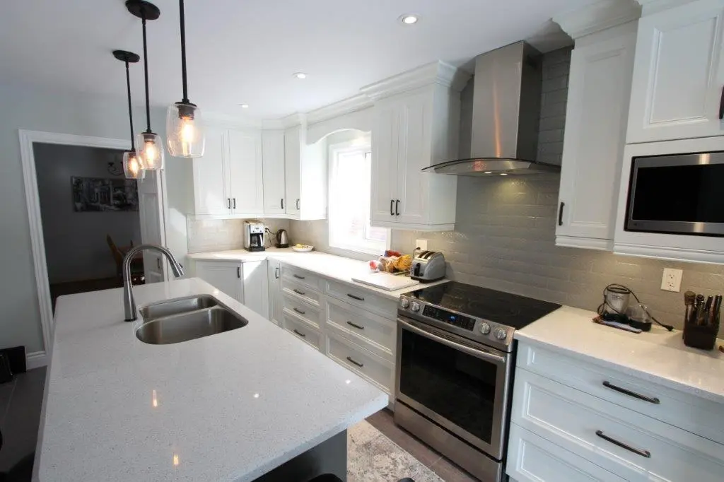 Modern kitchen with white cabinets, a large island with double sink, stainless steel oven and stovetop with a range hood, three hanging pendant lights, and various small appliances on the countertops under natural and recessed lighting.
