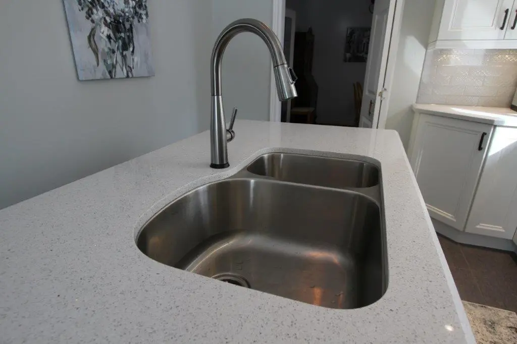 Modern kitchen countertop with a double stainless steel sink and a curved metal faucet, with white cabinets and a painting on the wall in the background