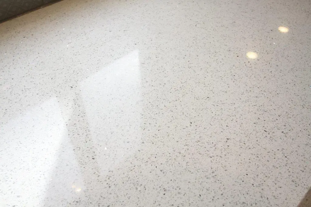 Close-up of a smooth, light-colored terrazzo surface with small dark speckles and reflections from overhead lights and a window.