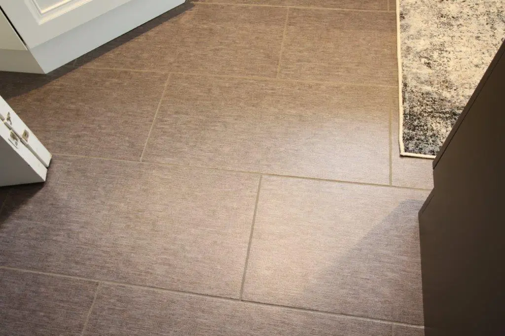 Close-up view of a tiled floor with a brownish-gray texture, a portion of a white cabinet in the top left corner, and part of a patterned rug in the top right corner.