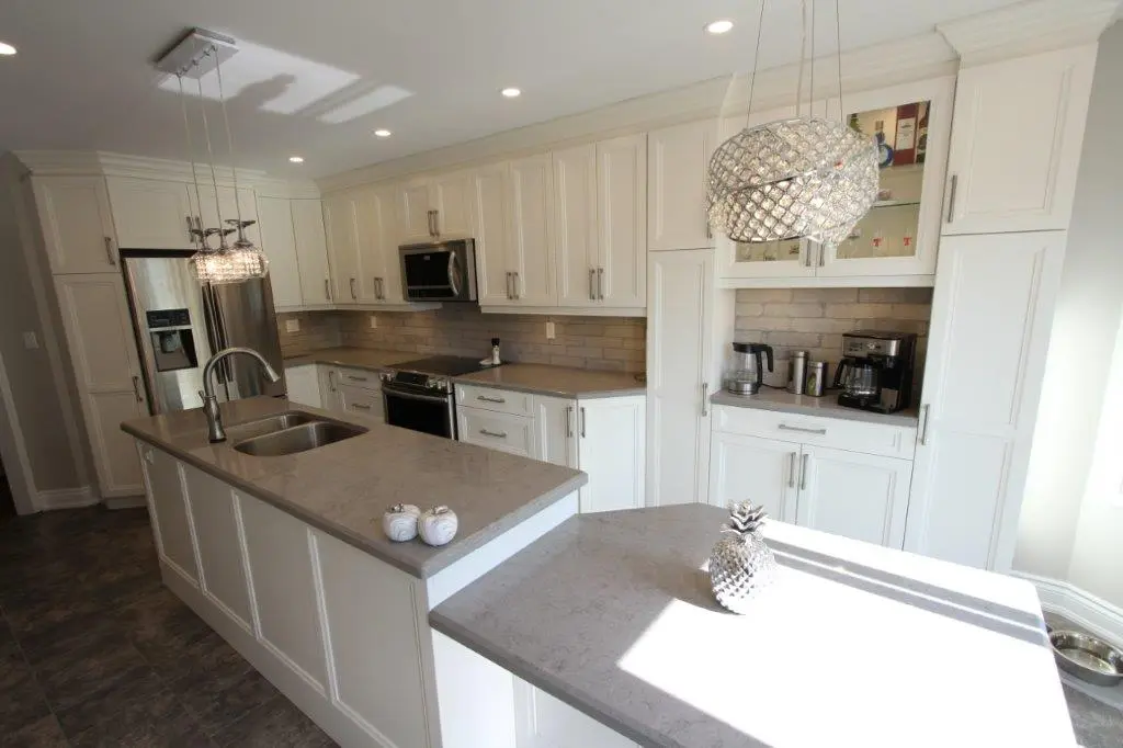 Modern kitchen with white cabinetry and gray countertops, featuring a large island with a built-in sink, stainless steel appliances, two crystal pendant lights, and a decorative silver pineapple on the counter in bright sunlight.