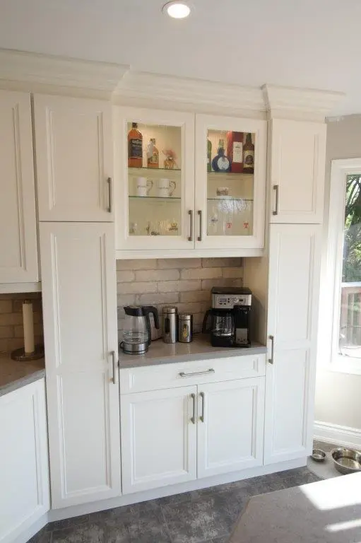 White kitchen cabinets with glass doors on the upper central section displaying bottles and mugs, countertop with a coffee maker, electric kettle, and a stainless steel travel mug, beige brick backsplash, and a window letting in natural light on the right side.