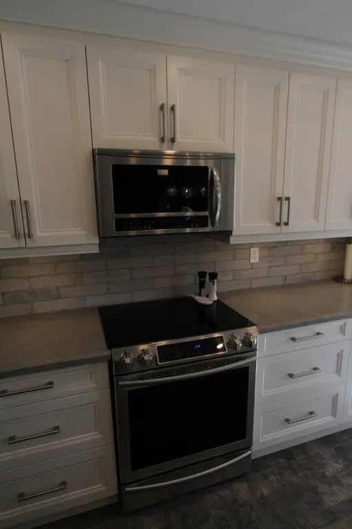 Modern kitchen with white cabinets, a stainless steel microwave mounted above a matching stainless steel electric stove with a smooth black cooktop, gray countertop, and beige brick-style backsplash.
